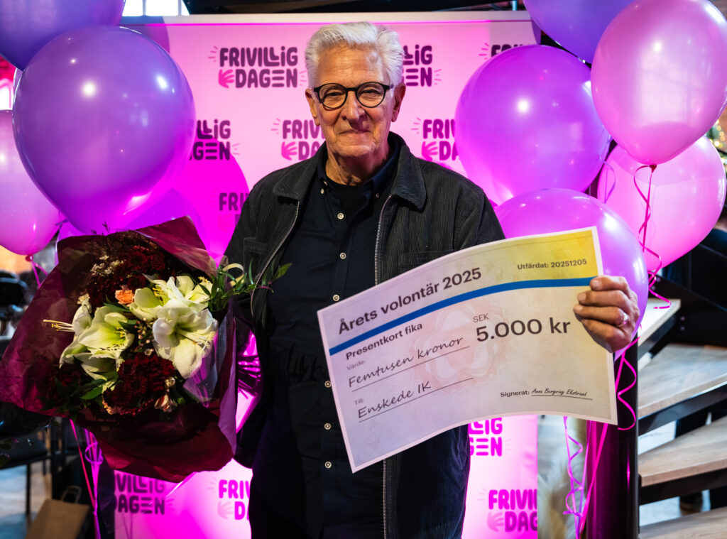 Äldre man håller blommor och en stor check framför en bakgrund med texten Frivilligdagen och ballonger.